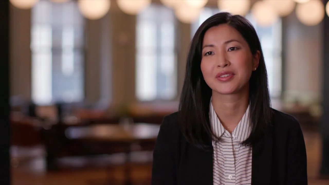 Woman with long dark hair speaking in a softly lit room with blurred background windows.