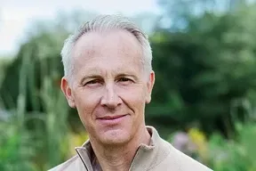 Smiling middle-aged man with short gray hair standing outdoors with greenery in the background.