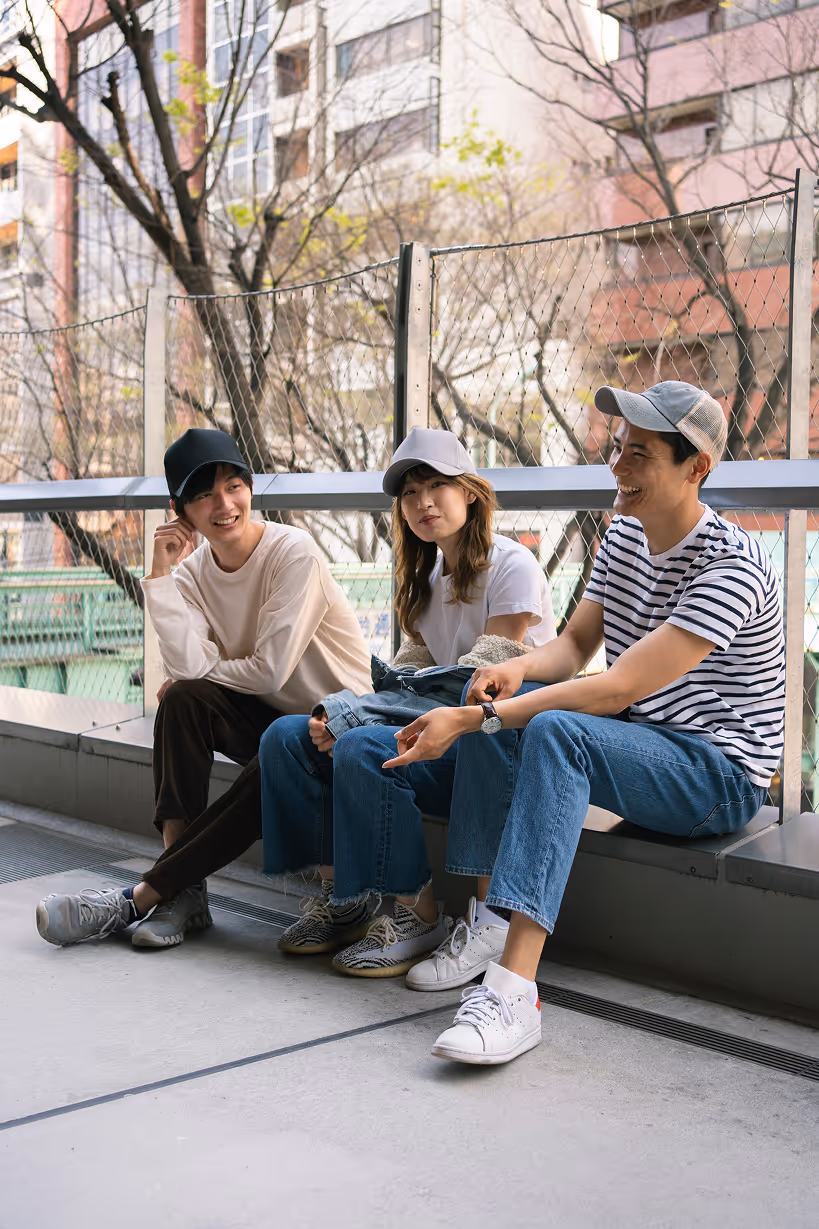 Three young people wearing caps sitting and chatting on an outdoor bench with buildings and trees in the background.
