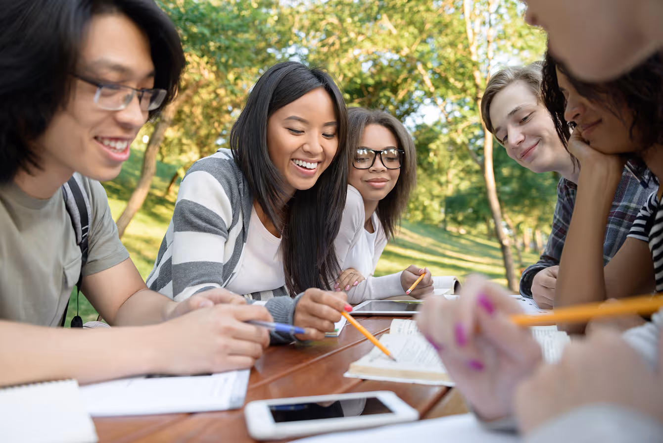 Group of diverse young adults studying together outdoors at a wooden table with notebooks, pencils, and a smartphone.