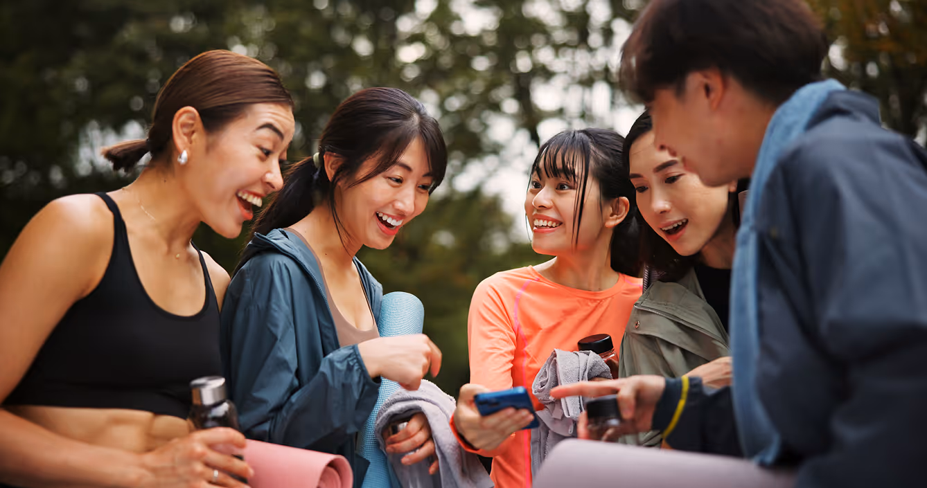 Group of young adults in athletic wear smiling and looking at a phone outdoors with yoga mats and water bottles.