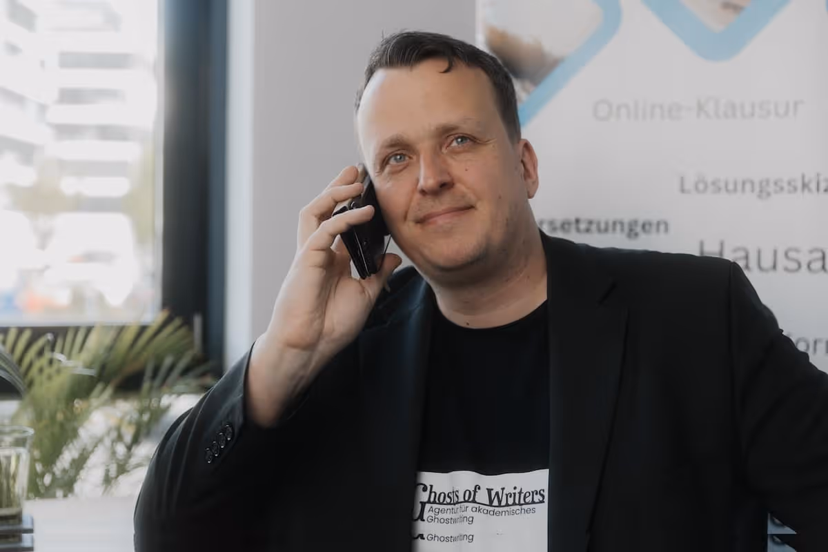 Mann im schwarzen Blazer telefoniert lächelnd in einem hellen Raum mit Pflanzen im Hintergrund.