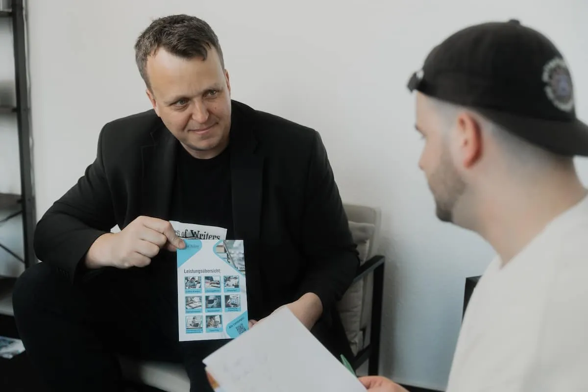 Zwei Männer sitzen in einem Büro, einer zeigt dem anderen ein Flyer mit einer Leistungsübersicht.