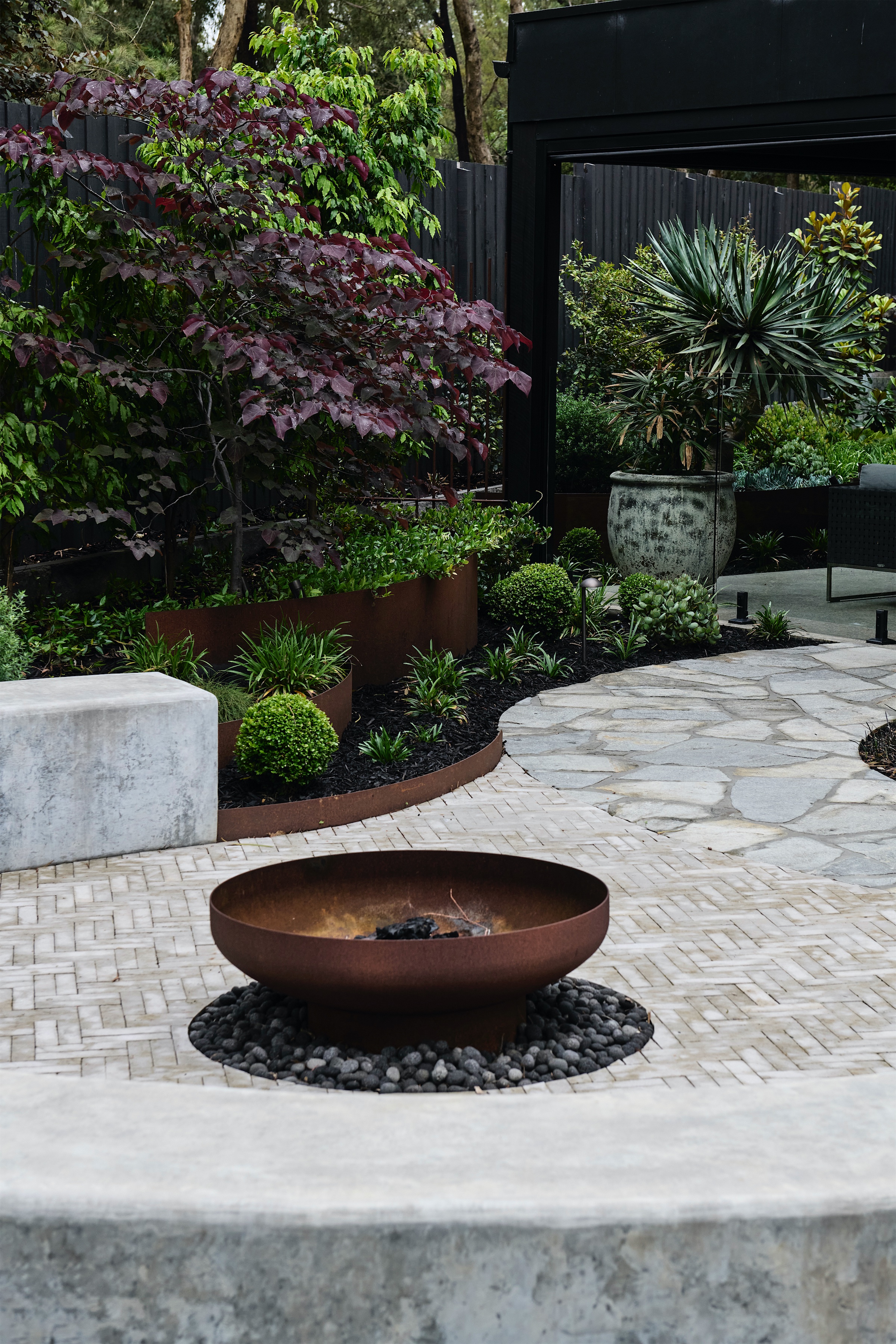 rusted steel fire pit on herringbone paving with a curved concrete bench, opus 2, ashburton