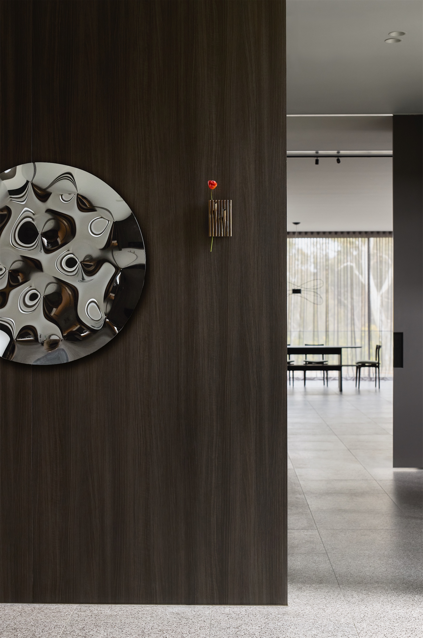 dark timber wall with a round polished-metal artwork and a red flower in a wall vase, opening to the dining room and trees beyond, opus 24, ashburton