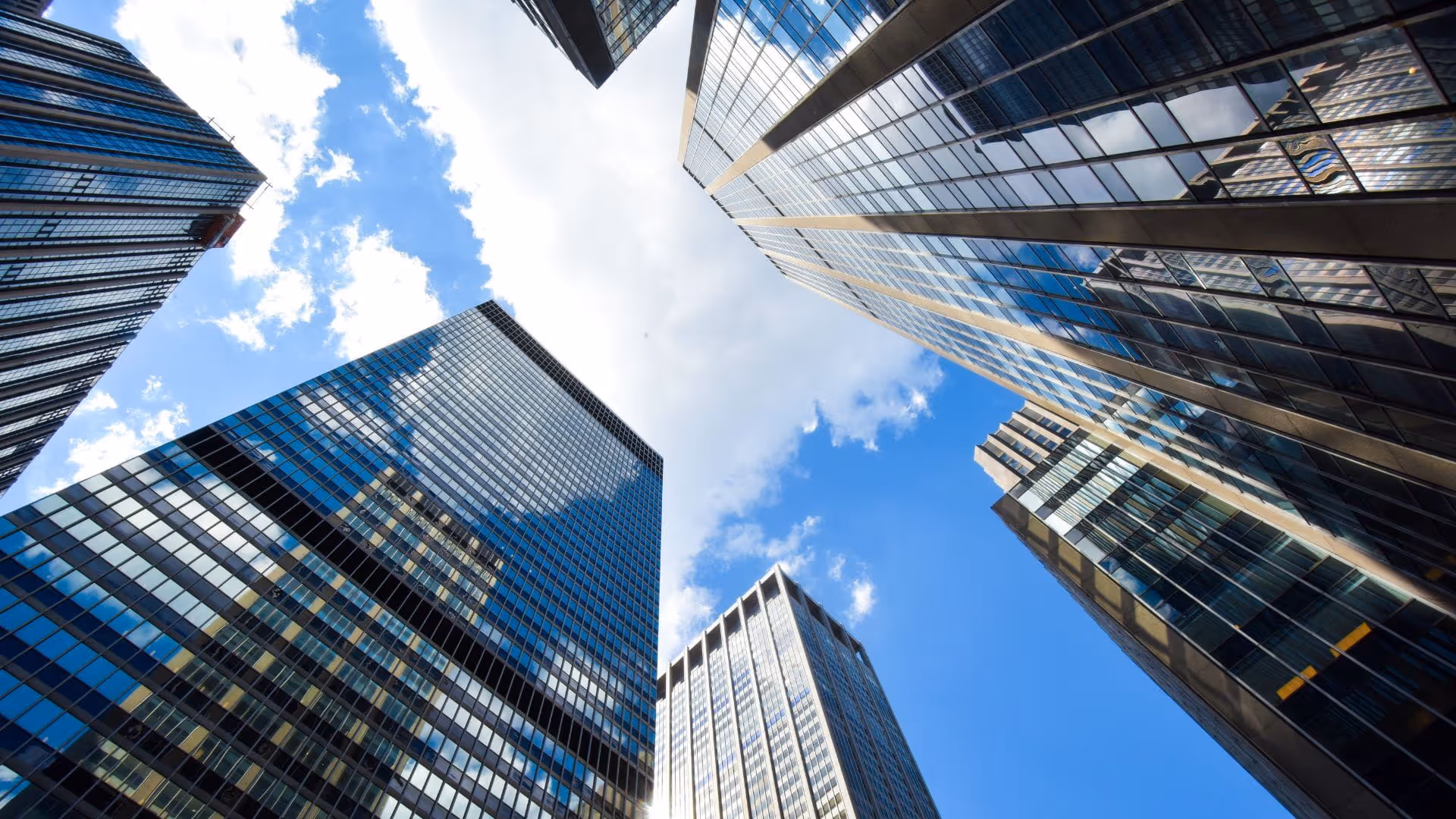 View looking up at tall glass skyscrapers against a blue sky with scattered clouds.