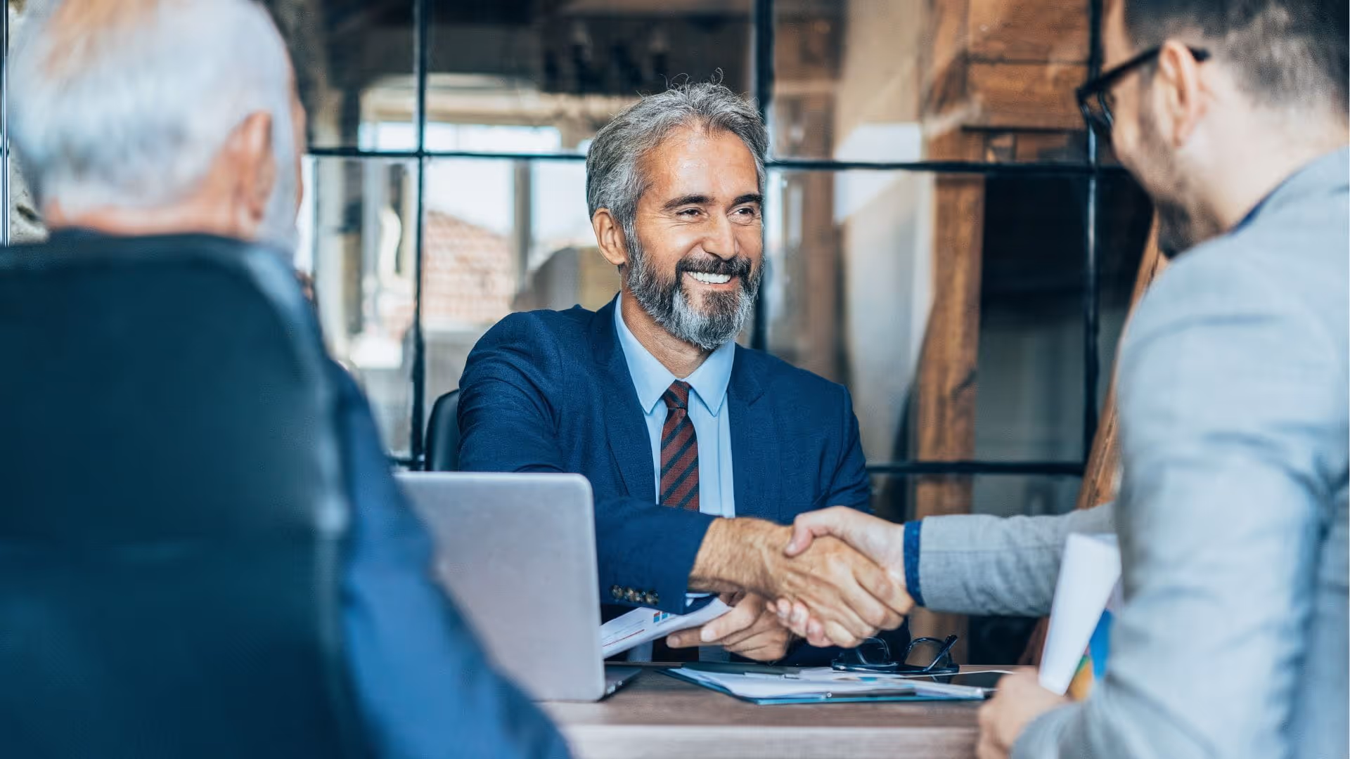 Smiling businessman in blue suit shaking hands with a colleague across a desk in an office.
