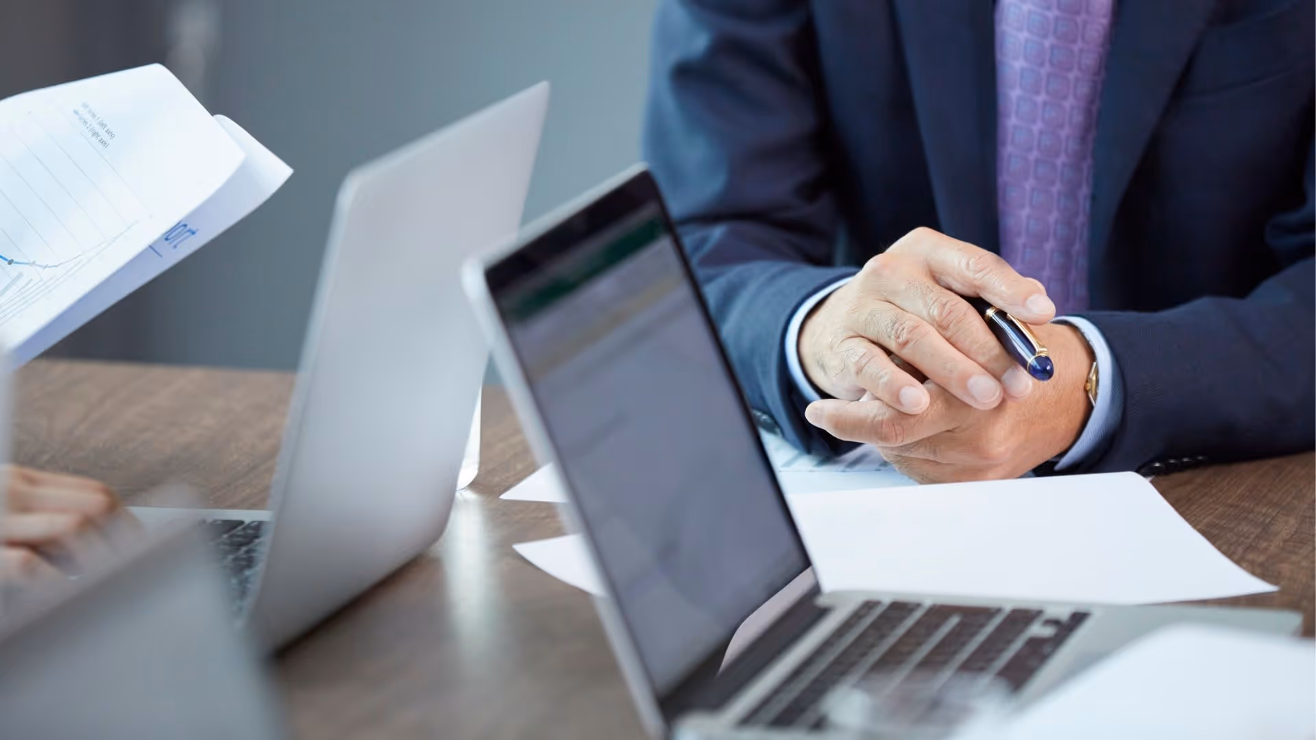 Two people working at a table with open laptops, documents, and one person holding a pen.