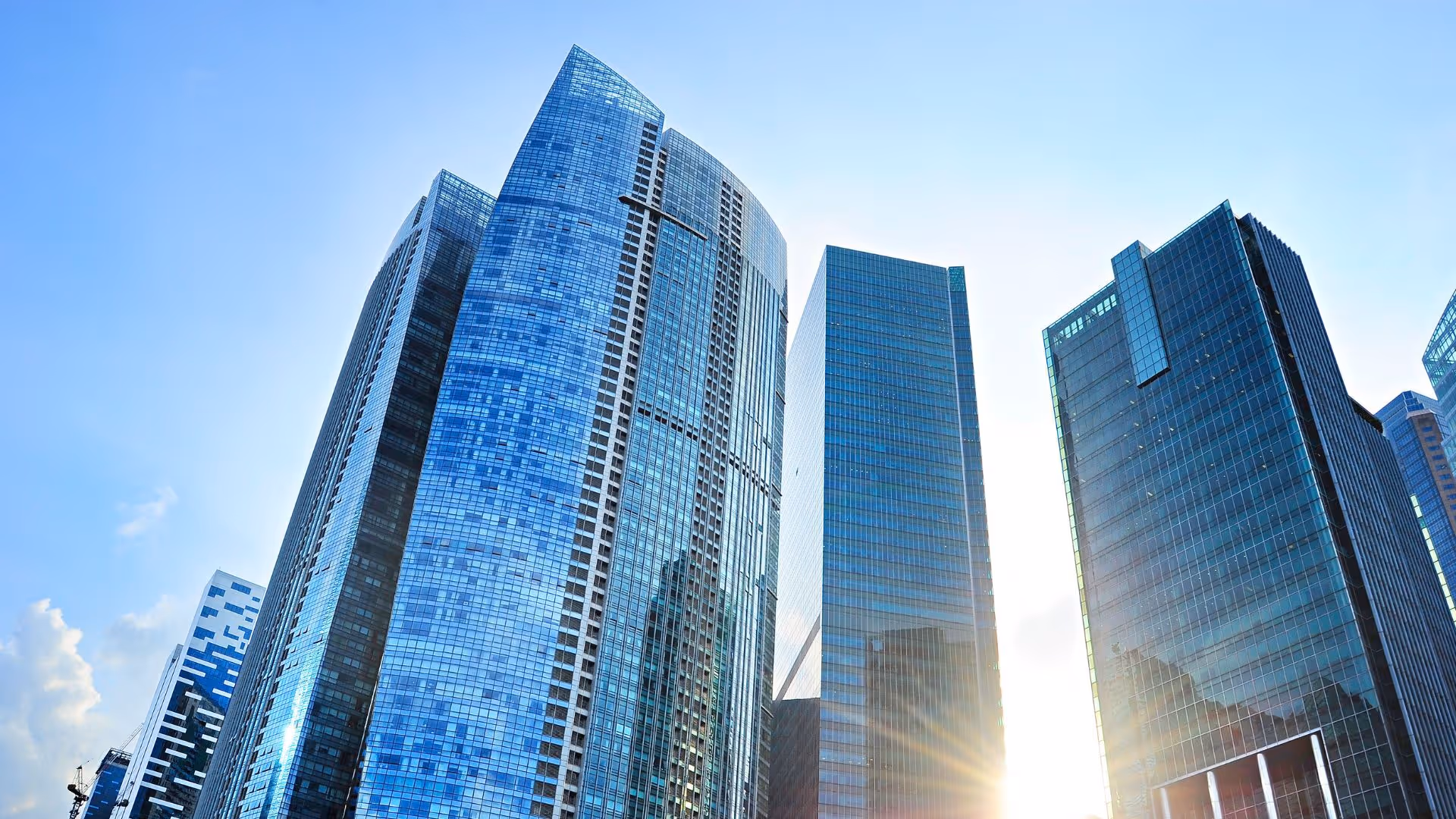 Sunlight shining through a cluster of tall modern glass skyscrapers against a clear blue sky.