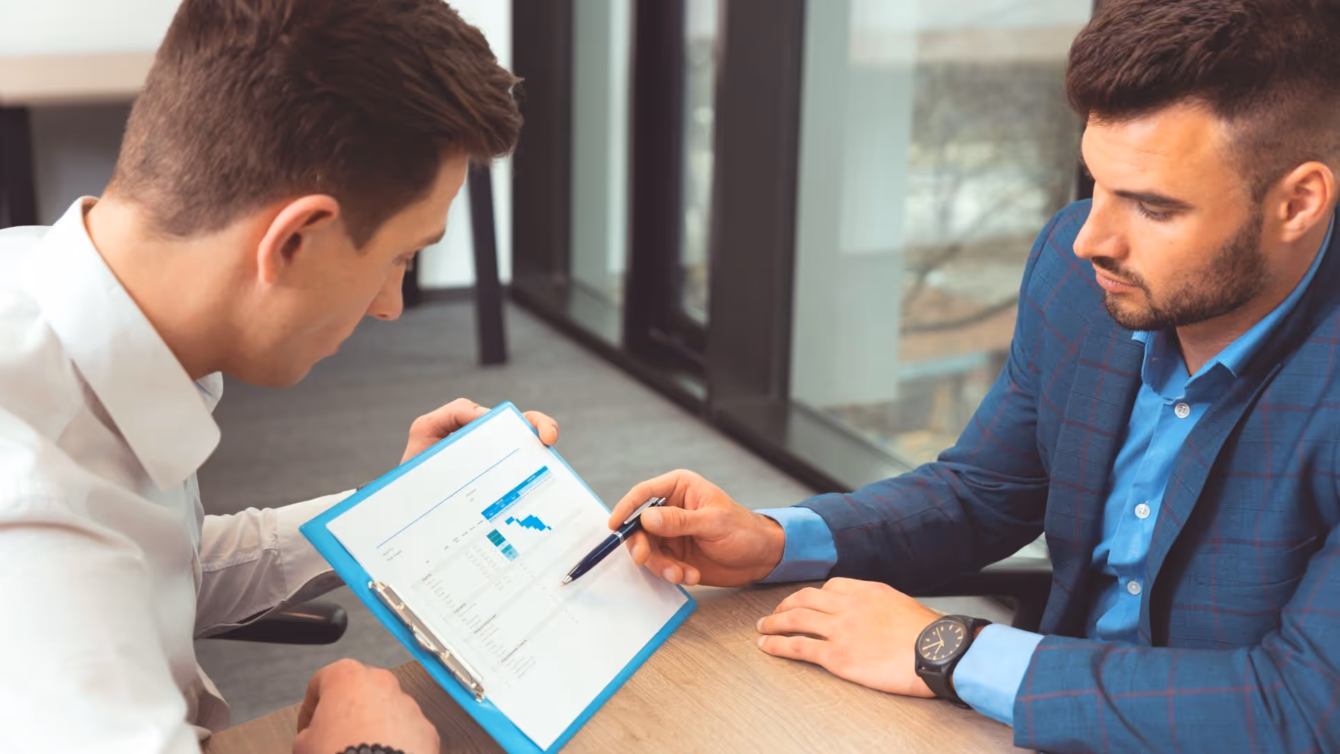 Two men in business attire reviewing a document with charts and data on a clipboard at a desk.