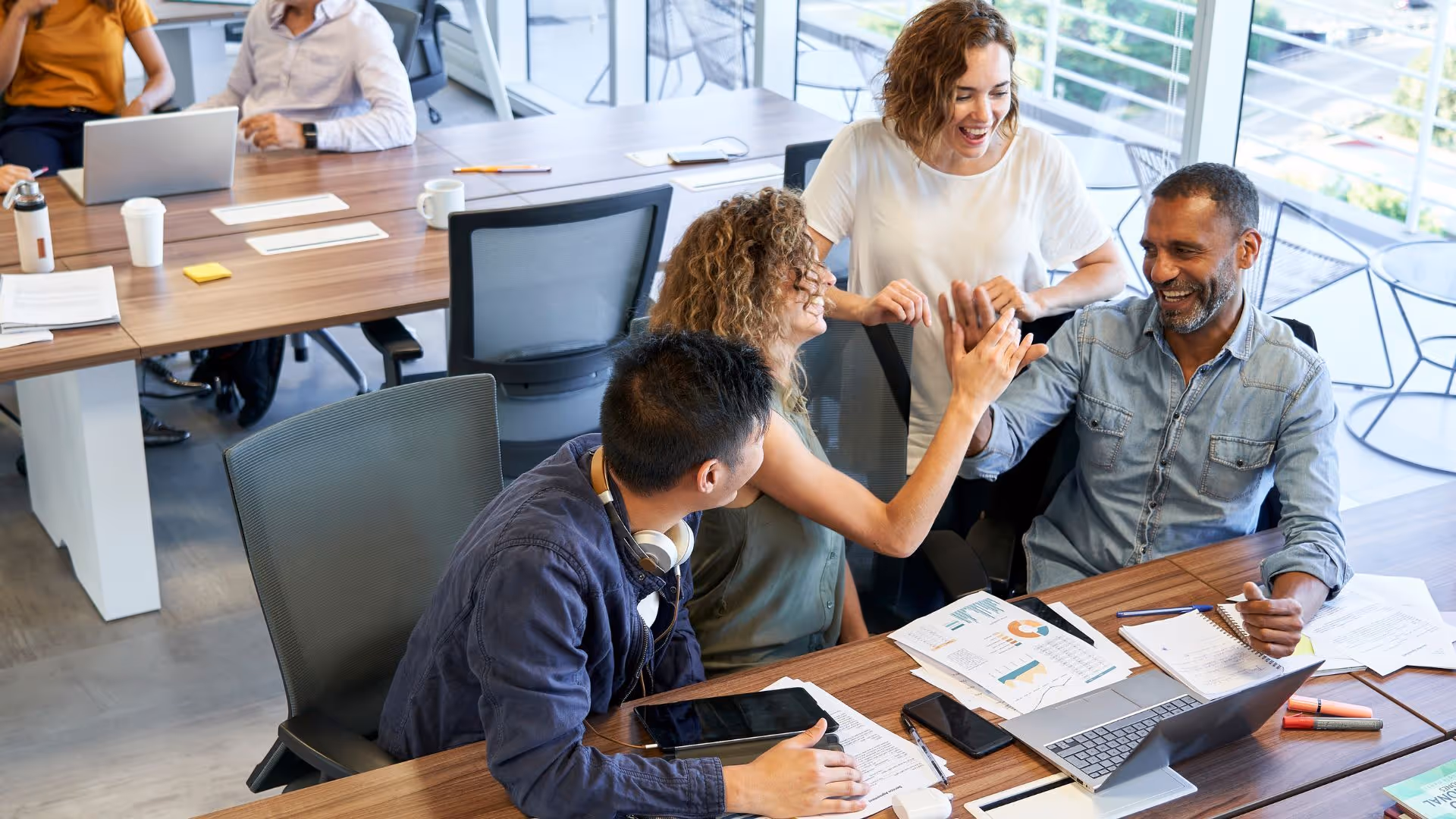 Three coworkers at a table in an office celebrating with a high-five, surrounded by papers, a laptop, and a smartphone.