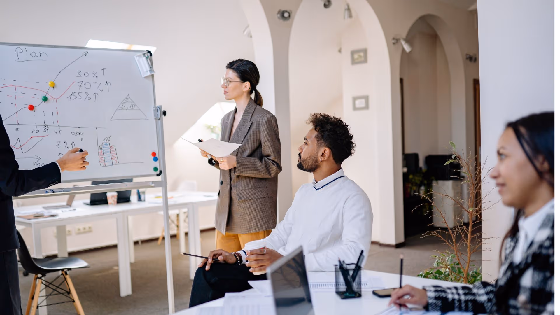 Three colleagues in an office meeting with one presenting a plan on a whiteboard while others listen and take notes.