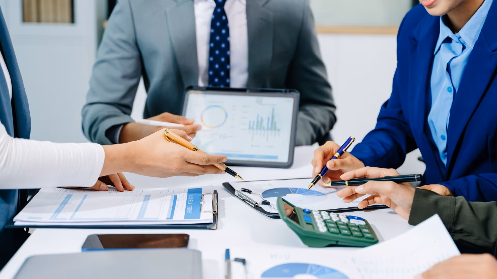 Business professionals collaborating at a table with documents, charts, a calculator, and a tablet displaying data graphs.