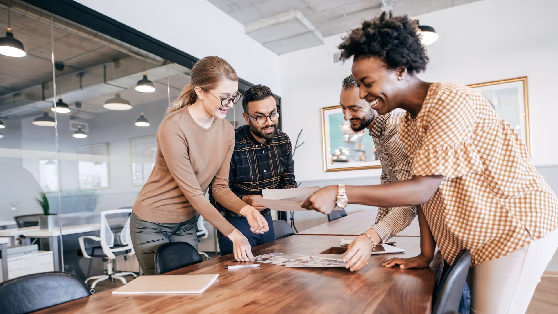 Four diverse colleagues smiling and reviewing papers together around a wooden conference table in a modern office.