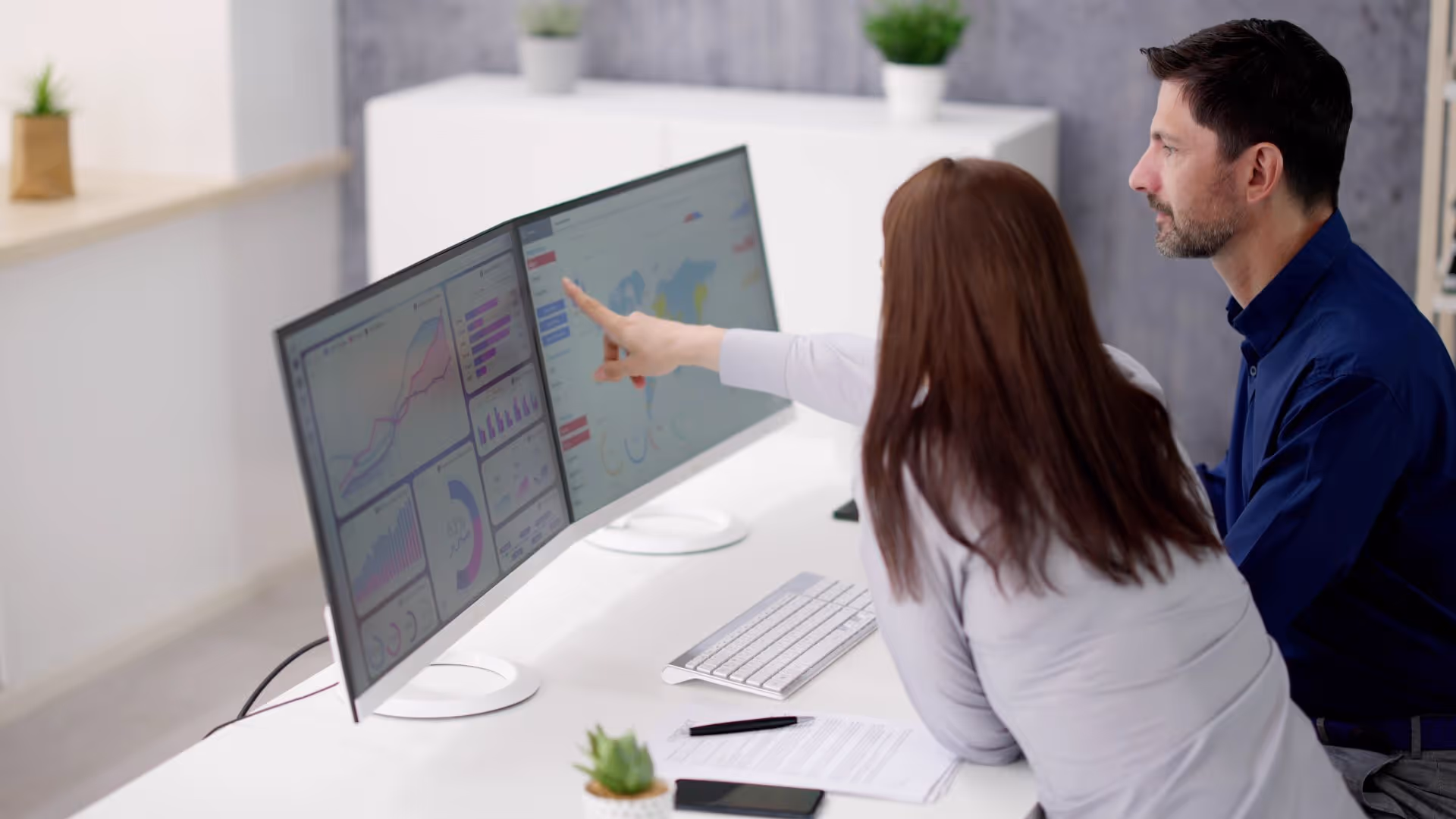 Two colleagues analyzing data charts on dual computer monitors in a modern office.