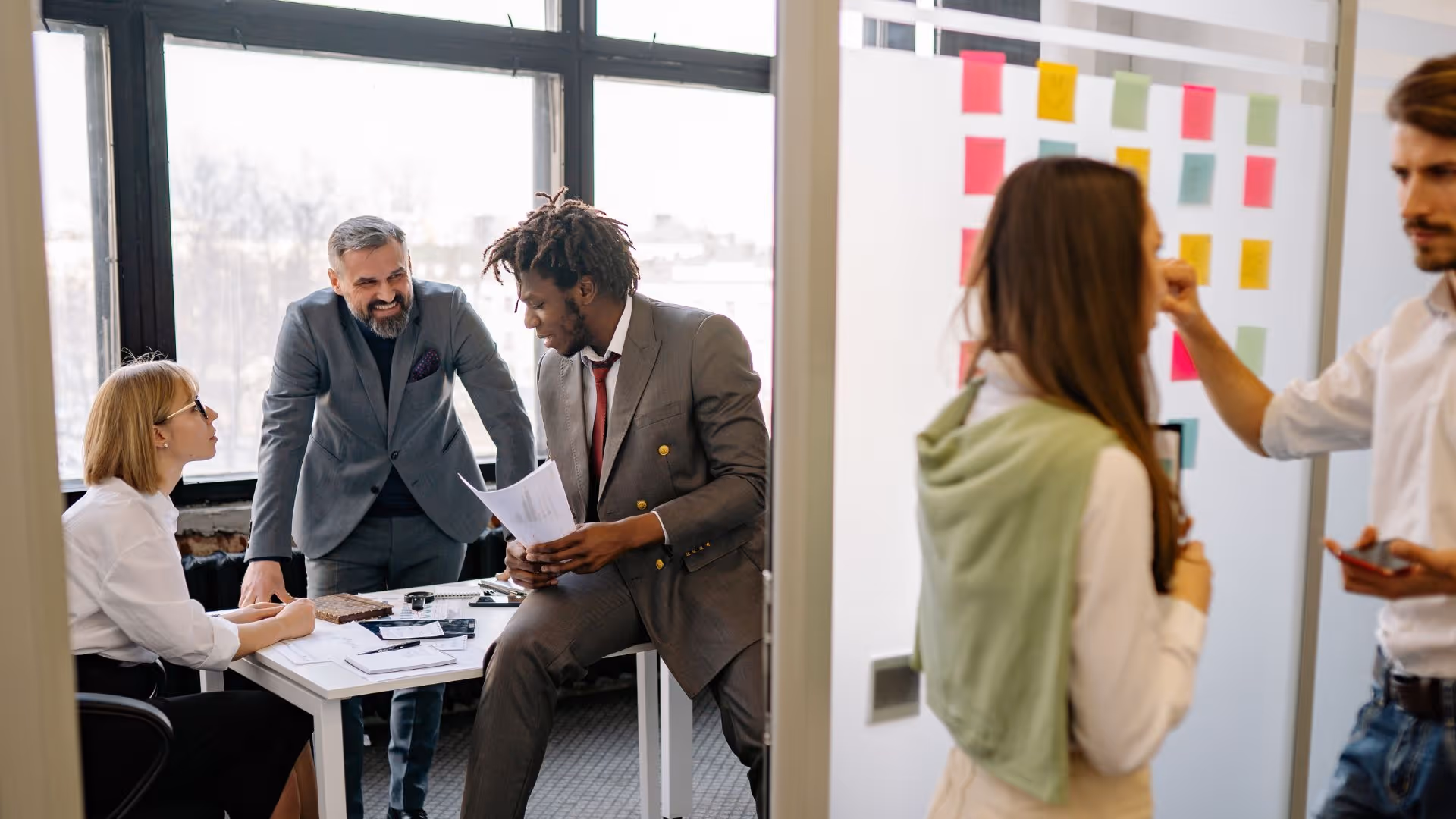 Five professionals collaborating in an office, with three discussing documents at a table and two others brainstorming near a whiteboard with colorful sticky notes.