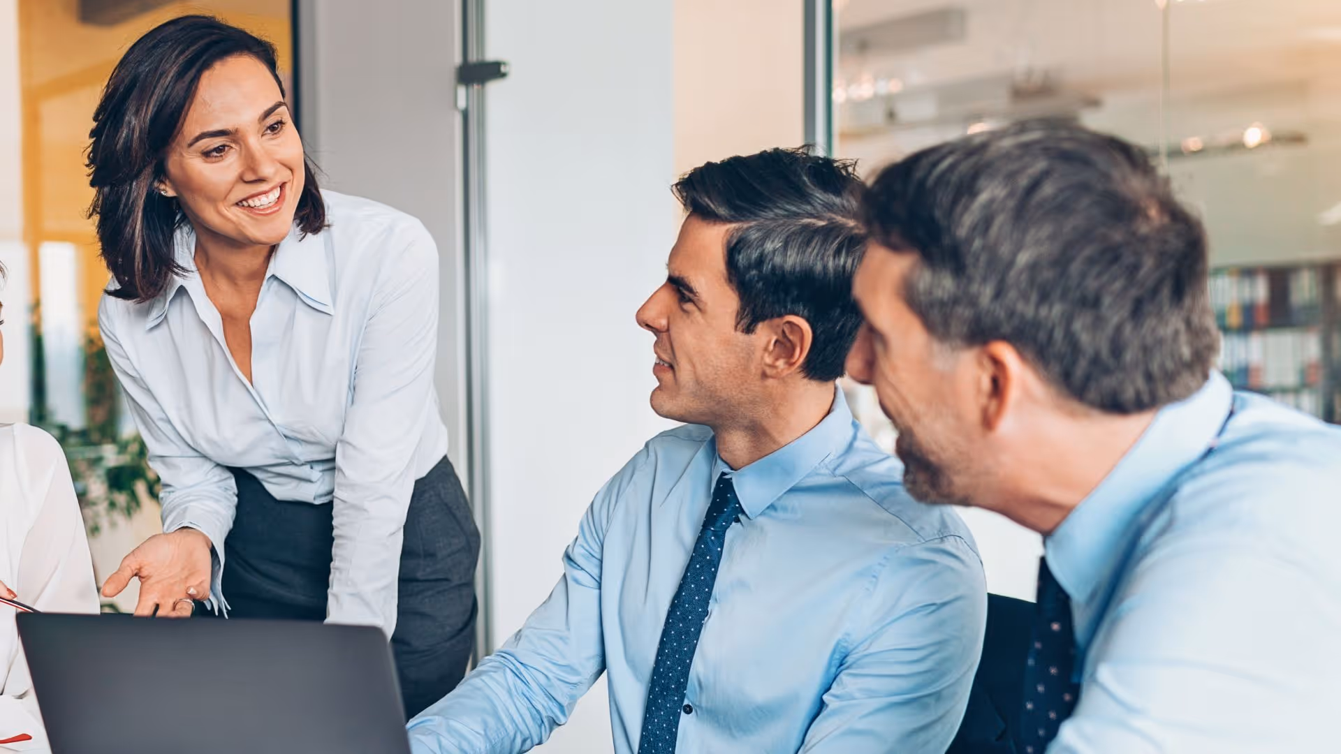 Businesswoman in white blouse smiling and talking to two men in light blue shirts and ties during a meeting.