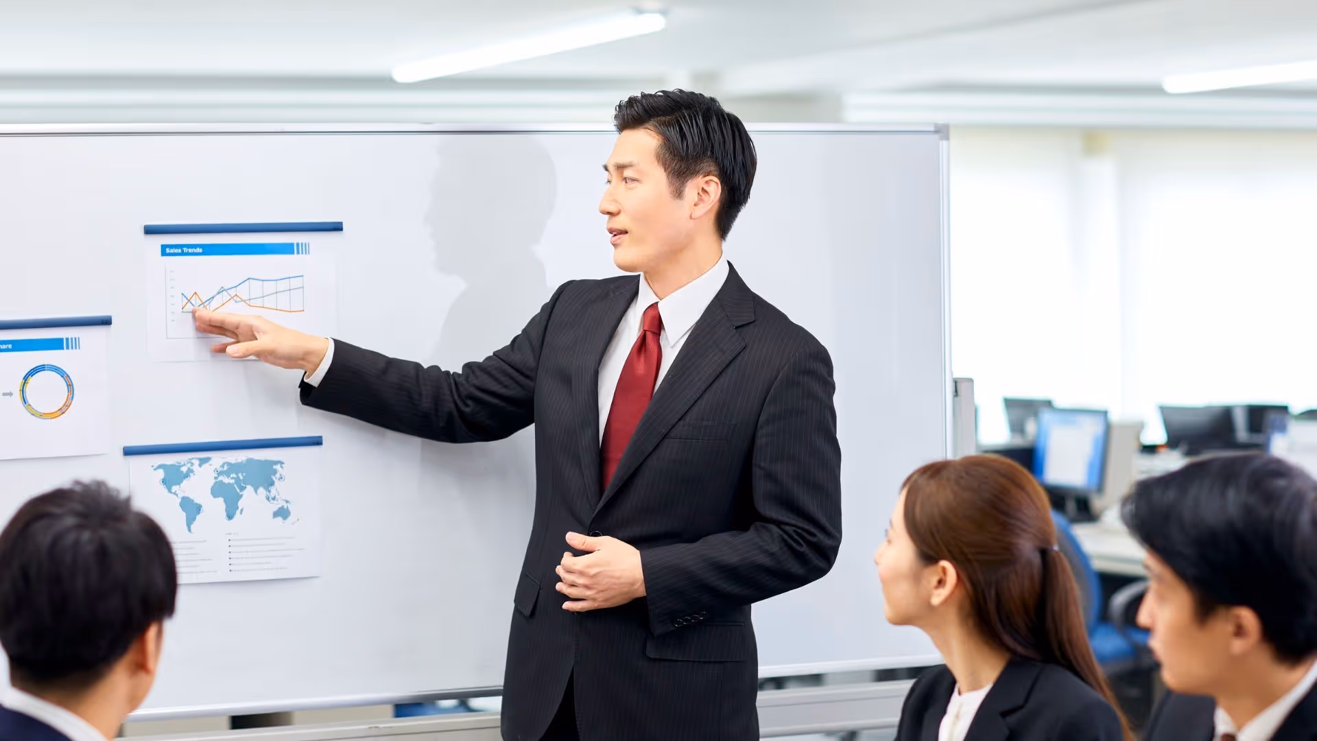 Businessman in a suit pointing at sales charts on a whiteboard during a meeting with colleagues.