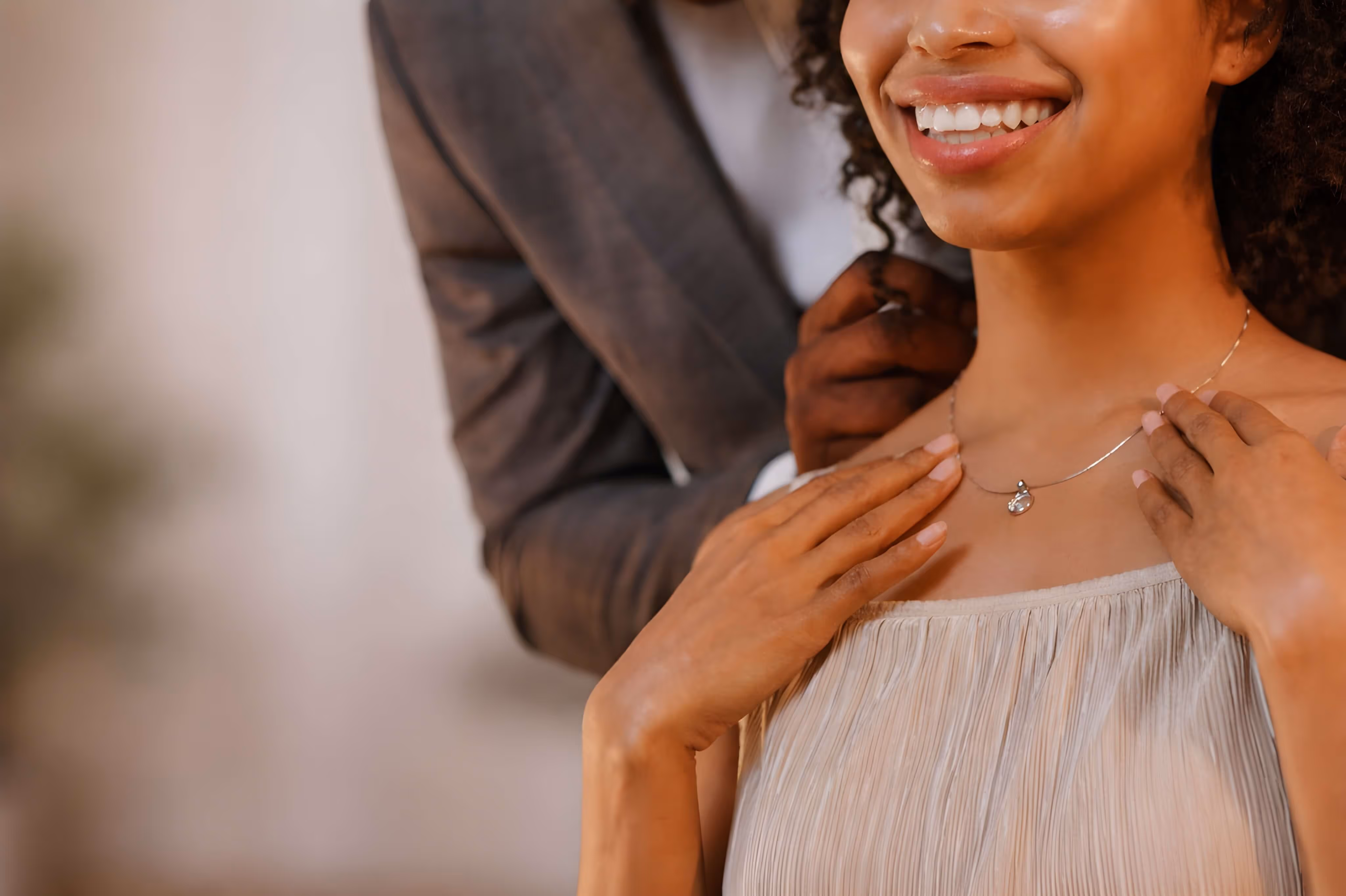 Close-up of a smiling woman wearing a delicate necklace while a person behind her clasps it around her neck.