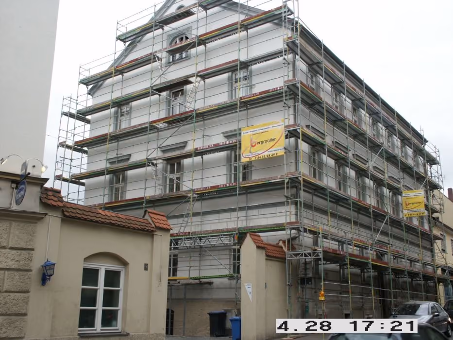 Ein mehrstöckiges Wohngebäude, das von Gerüstmatter umgeben ist, mit mehreren Fenstern und einem Tor mit Ziegelgedeck an der Vorderseite an einem bewölkten Tag.