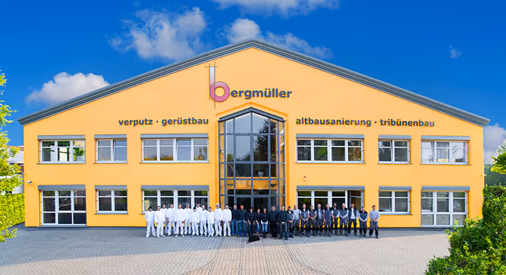 Gelbes zweistöckiges Firmengebäude mit vielen Fenstern und einem großen gläsernen Vordach im Zentrum, darüber das Firmenlogo 'bergmüller' und die Beschriftungen 'verputz · gerüstbau', 'altbausanierung · tribünenbau'. Vor dem Gebäude steht eine große Gruppe von Mitarbeitern in weißer, dunkler und grauer Arbeitskleidung auf gepflastertem Vorplatz unter blauem Himmel mit wenigen Wolken.