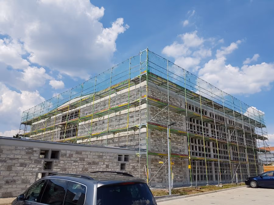 Ein großes rechteckiges Gebäude aus Stein mit Gerüst und Sicherheitsnetzen an den Wänden unter blauem Himmel mit einigen Wolken, im Vordergrund zwei parkende Autos.