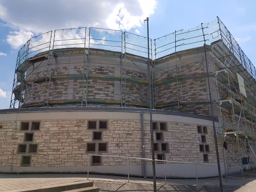 Gebäude mit Steinfassade, teilweise von Gerüsten umgeben für Renovierungsarbeiten unter blauem Himmel mit Wolken.