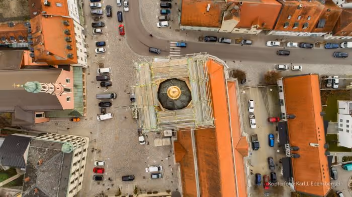 Luftaufnahme einer Innenstadt mit Kopfsteinpflaster, umgeben von Gebäuden mit roten Dächern, darunter eine Kirche mit grünem Turm und ein Gebäude mit einem Kuppeldach in der Mitte.