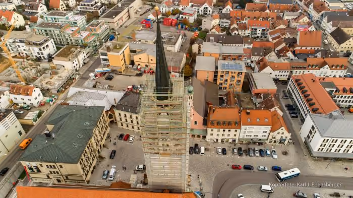 Luftaufnahme eines Stadtkerns mit einem hohen Kirchturm in der Bildmitte, der mit einem Gerüst zur Renovierung umgeben ist, rundherum zahlreiche Häuser mit roten Dächern und geparkten Autos auf Pflasterstraßen.