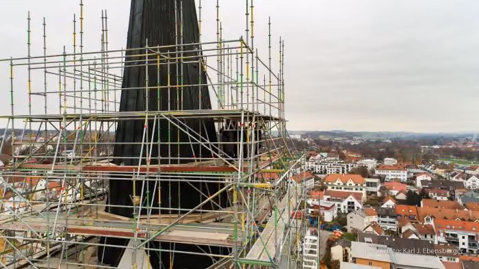 Turm einer Kirche oder eines Gebäudes, umgeben von Gerüstkonstruktionen für Renovierungsarbeiten, mit Blick auf ein Wohngebiet mit roten Dächern unter bewölktem Himmel.