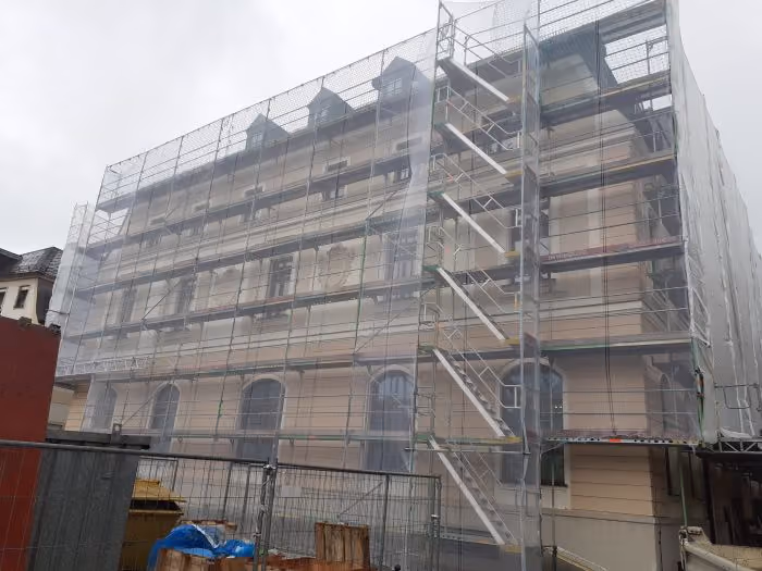 Mehrstöckiges Gebäude mit kleinen Dachgauben, eingehüllt in ein Gerüst mit Schutznetz, Baustelle mit Bauzaun und Baumaschinen im Vordergrund bei bewölktem Himmel.