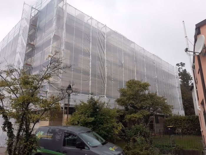 Großes Gebäude ist vollständig mit einem weißen Schutznetz für Renovierungsarbeiten bedeckt. Vor dem Gebäude stehen mehrere Bäume und ein dunkler Transporter mit der Aufschrift ‚Peltonenser‘ auf der Seite. Es ist bewölkter Himmel.