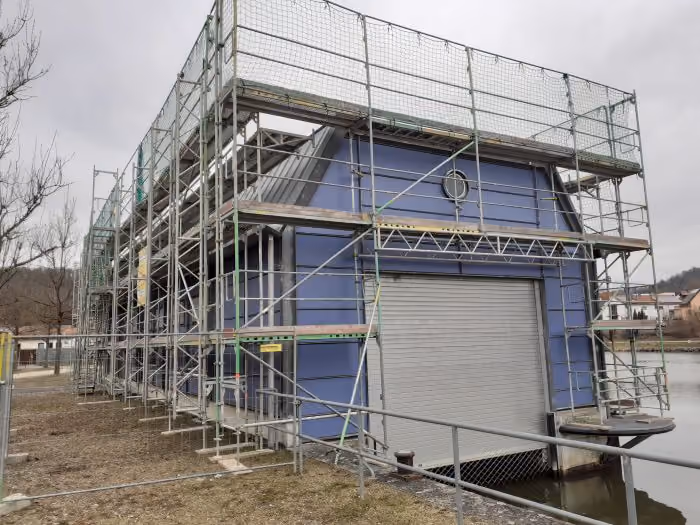 Gebäude in blauer Verkleidung, das von einem Metalldach und einem Stahlgerüst umgeben ist, steht am Ufer eines Gewässers an einem trüben Tag.