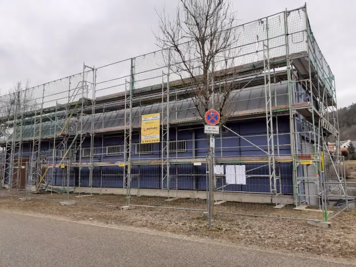 Ein mehrstöckiges Gebäude mit blauen Wänden steht im Umbau, umgeben von einem Gerüst aus Metallstangen, zwei gelben Warnschildern und einem Verkehrszeichen für Parkverbot vor grauem Himmel.