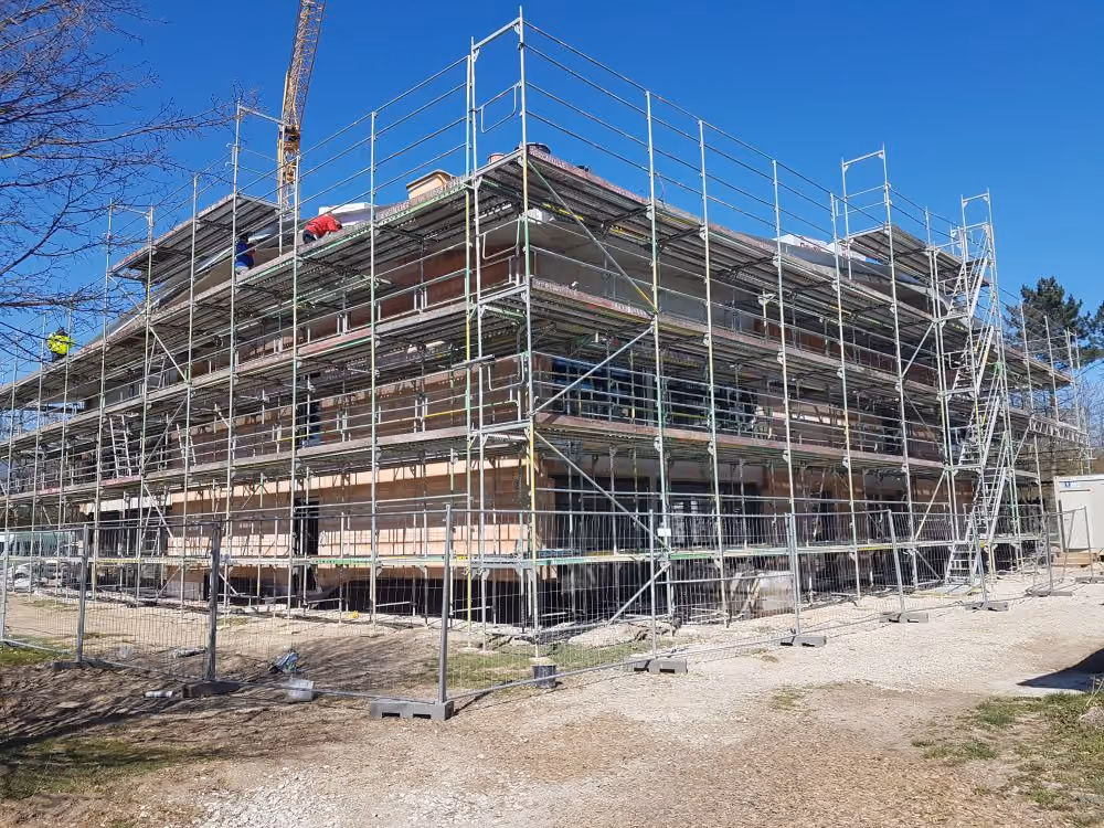 Eine große Baustelle mit einem mehrstöckigen Gebäude, das von Gerüsten umgeben ist, unter klarem blauem Himmel.