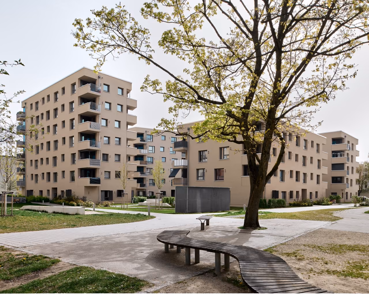 Wohnanlage mit modernen, beigefarbenen mehrstöckigen Gebäuden, Baum und geschwungener Holzbank im Hof.