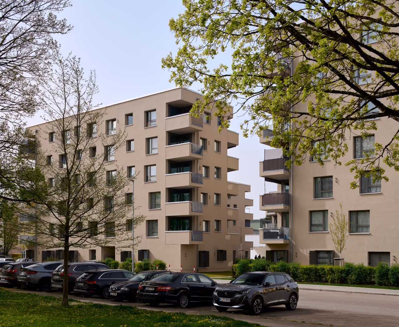 Mehrstöckige Wohnhäuser mit Balkonen, Autos parken am Straßenrand, Bäume mit frischem Grün.