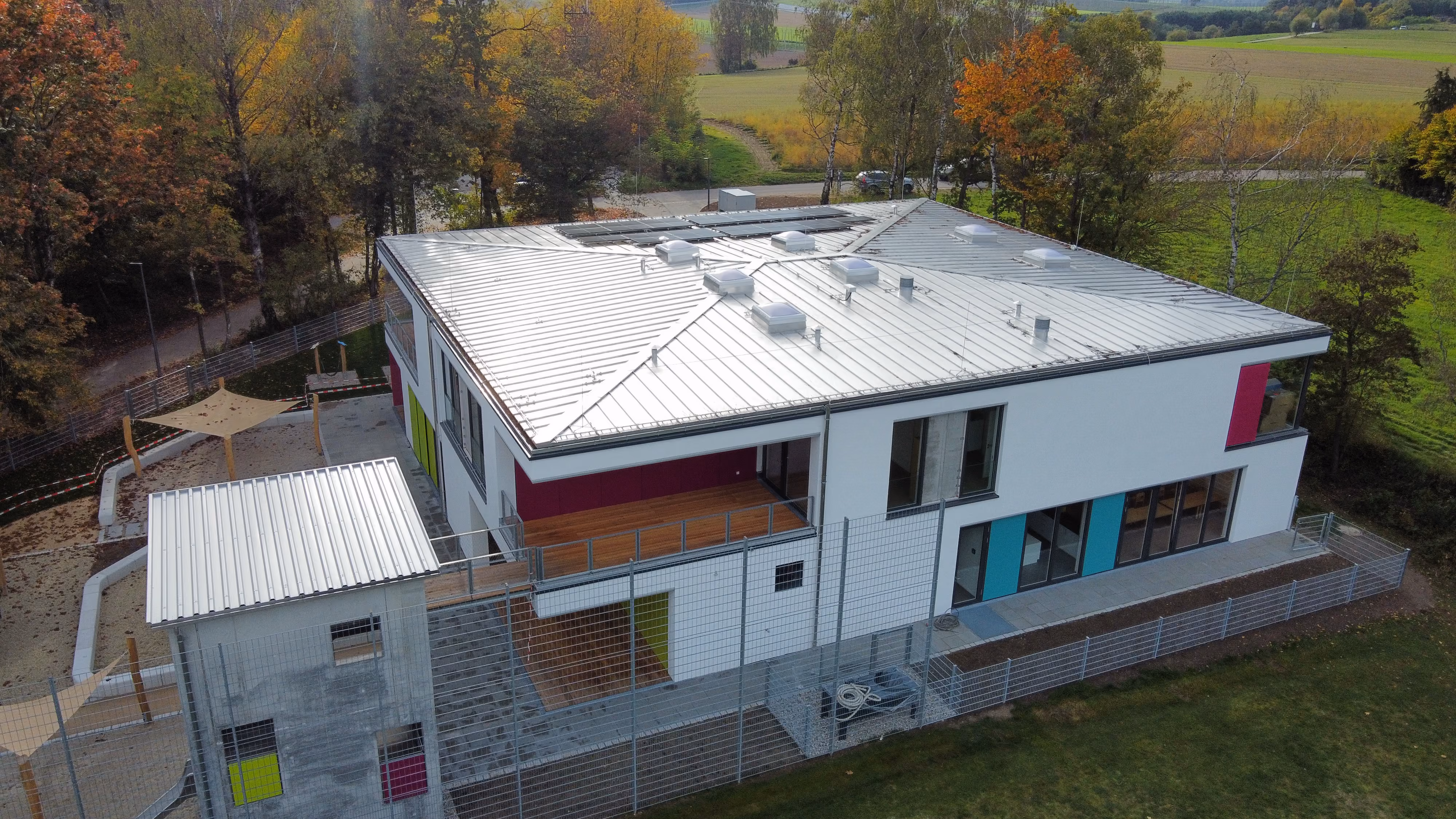 Modern zweistöckiges Gebäude mit weißer Fassade, Metallflachdach und bunten Akzenten, umgeben von Bäumen
