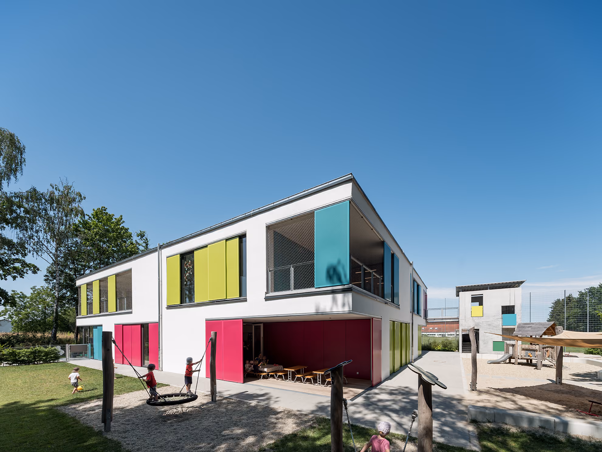Modern kindergarten building with colorful panels and children playing outside on swings and grass.