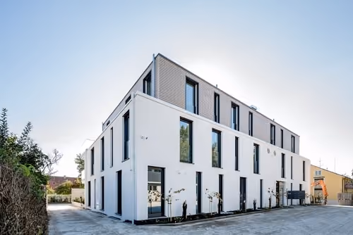 Modernes, mehrstöckiges Gebäude mit weißen und grauen Fassaden, vielen großen Fenstern und einer Einfahrt daneben unter einem klaren Himmel.