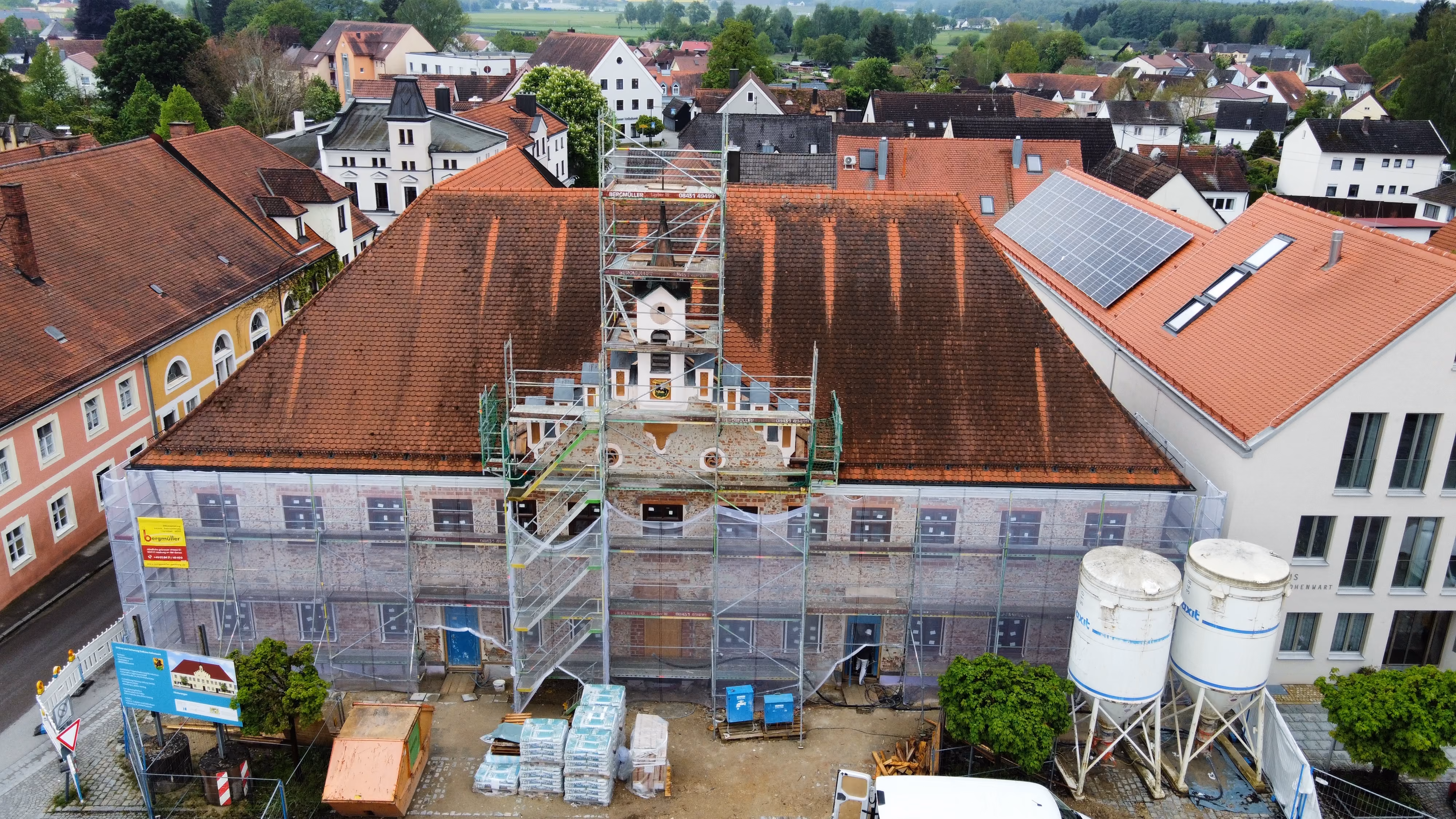 Blick auf ein historisches Gebäude mit roten Ziegeldächern in einer kleinen Stadt, das von Gerüsten und Baugerüsten umgeben ist, während Renovierungsarbeiten stattfinden. Stapel von Baumaterialien, zwei große Silos und Fahrzeuge sind vor dem Gebäude zu sehen, umgeben von typischer Wohnbebauung und grüner Landschaft im Hintergrund.