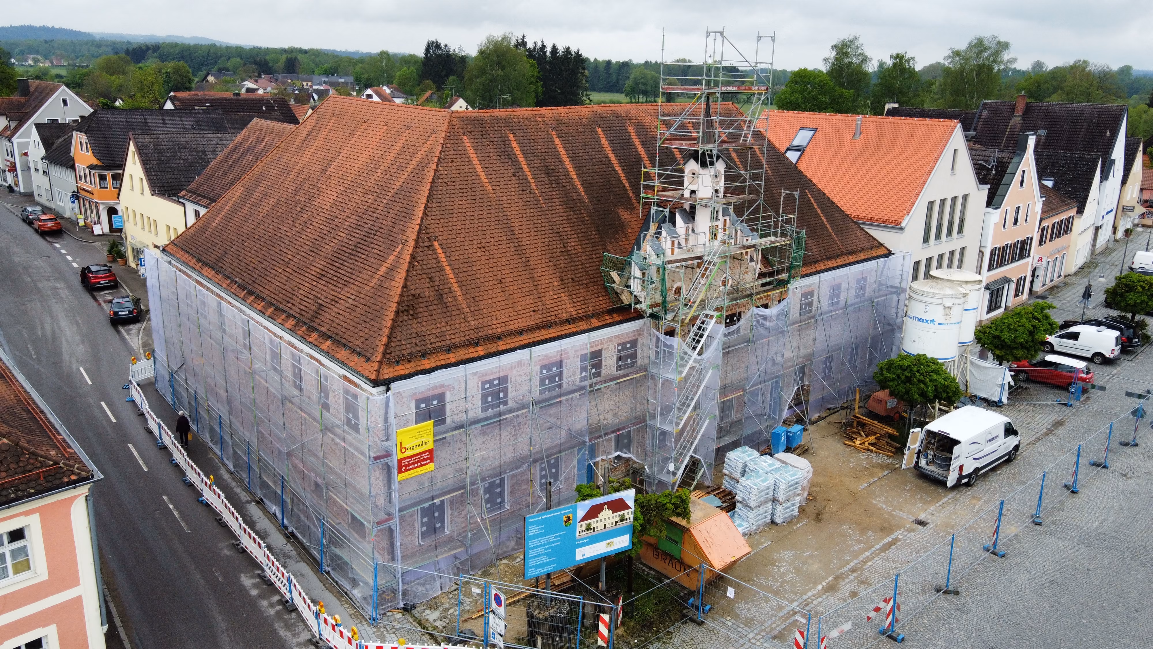 Ein historisches Gebäude in einer kleinen Stadt wird renoviert, mit einem großen Gerüst und Abdeckungen an den Wänden, umgeben von Straßen und parkenden Fahrzeugen.