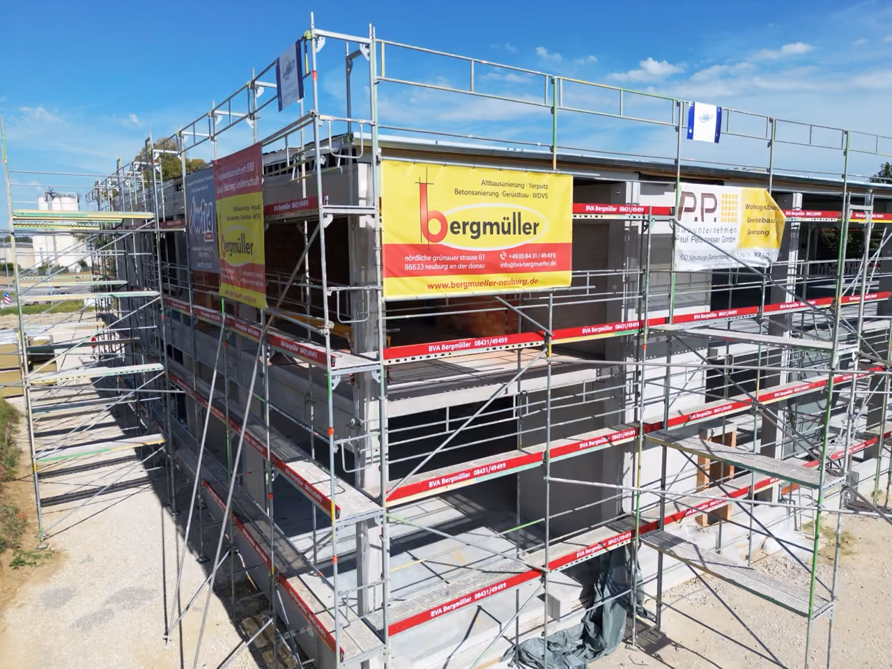 Neubau mit Gerüst und Banners verschiedener Baufirmen bei sonnigem Wetter.