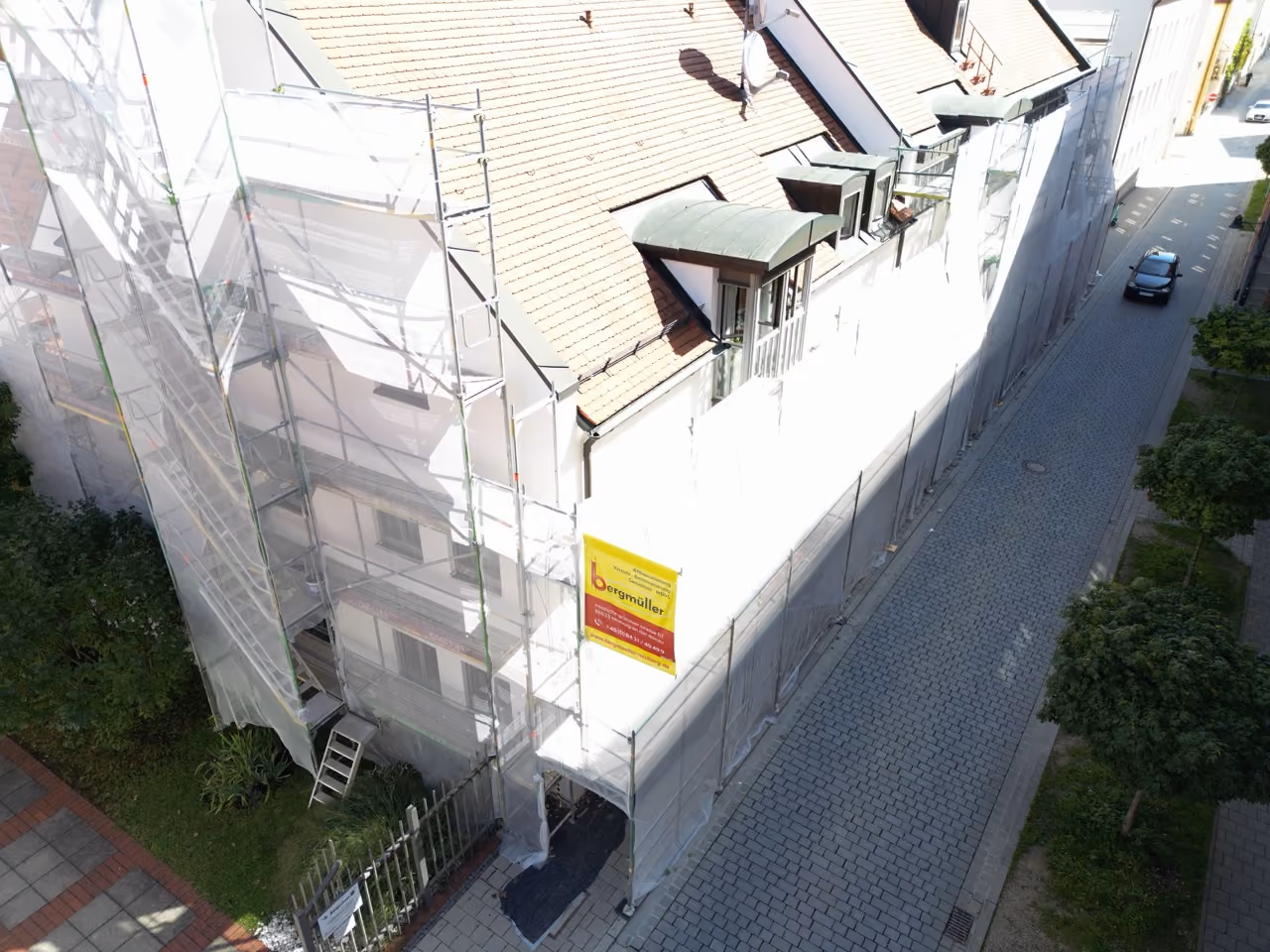 Baugerüst mit Schutzplane bedeckt eine Gebäudefassade an einer gepflasterten Straße mit einem parkenden Auto, umgeben von Bäumen und Grünflächen.