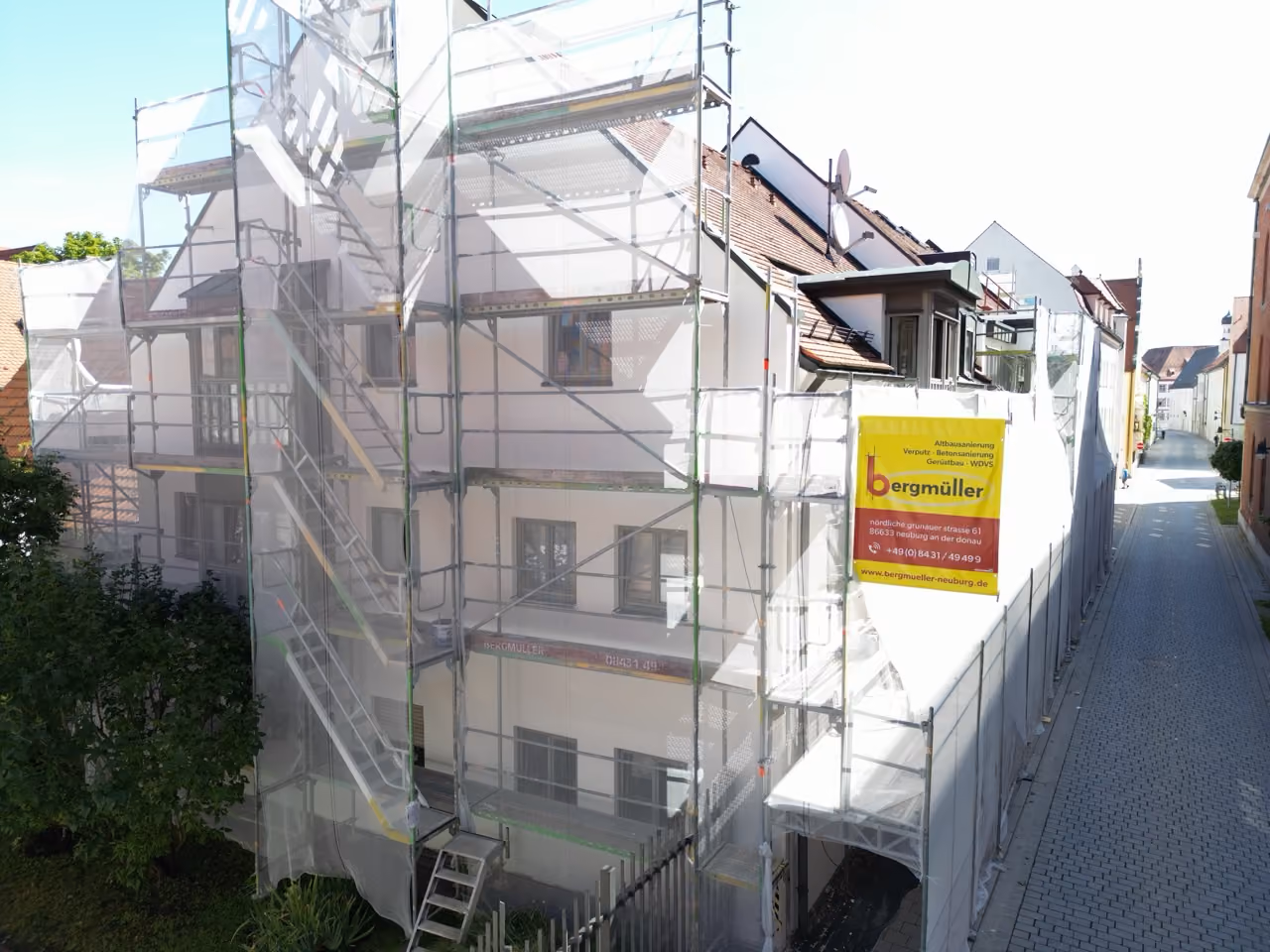 Ein mehrstöckiges Wohnhaus in einer engen Gasse ist für Renovierungsarbeiten mit Gerüst und weißem Schutznetz versehen, mit einem gelben Schild der Firma Bergmüller für Abbruch- und Sanierungsarbeiten.