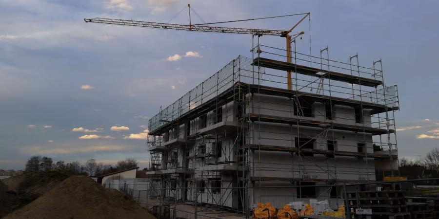 Mehrstöckiges Gebäude im Rohbau mit Baugerüsten an den Seiten und einem Kran darüber, bei bewölktem Himmel im Hintergrund.