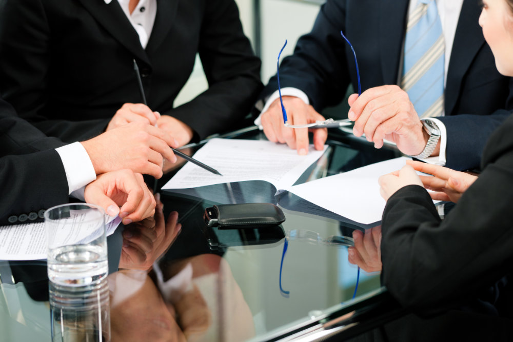 Quatre personnes en costume discutent autour d'une table en verre, examinant des documents et tenant un stylo et des lunettes.