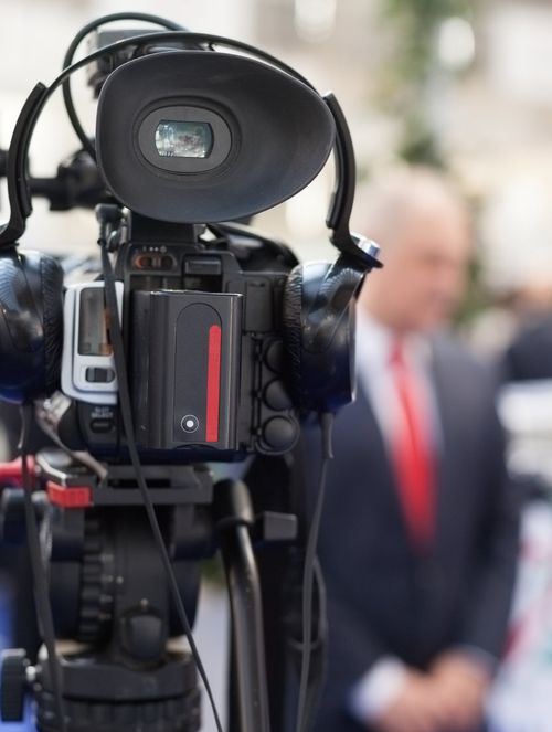 Caméra vidéo professionnelle avec casque d'écoute fixé, en premier plan, filmant un homme flou en costume et cravate rouge à l'arrière-plan.