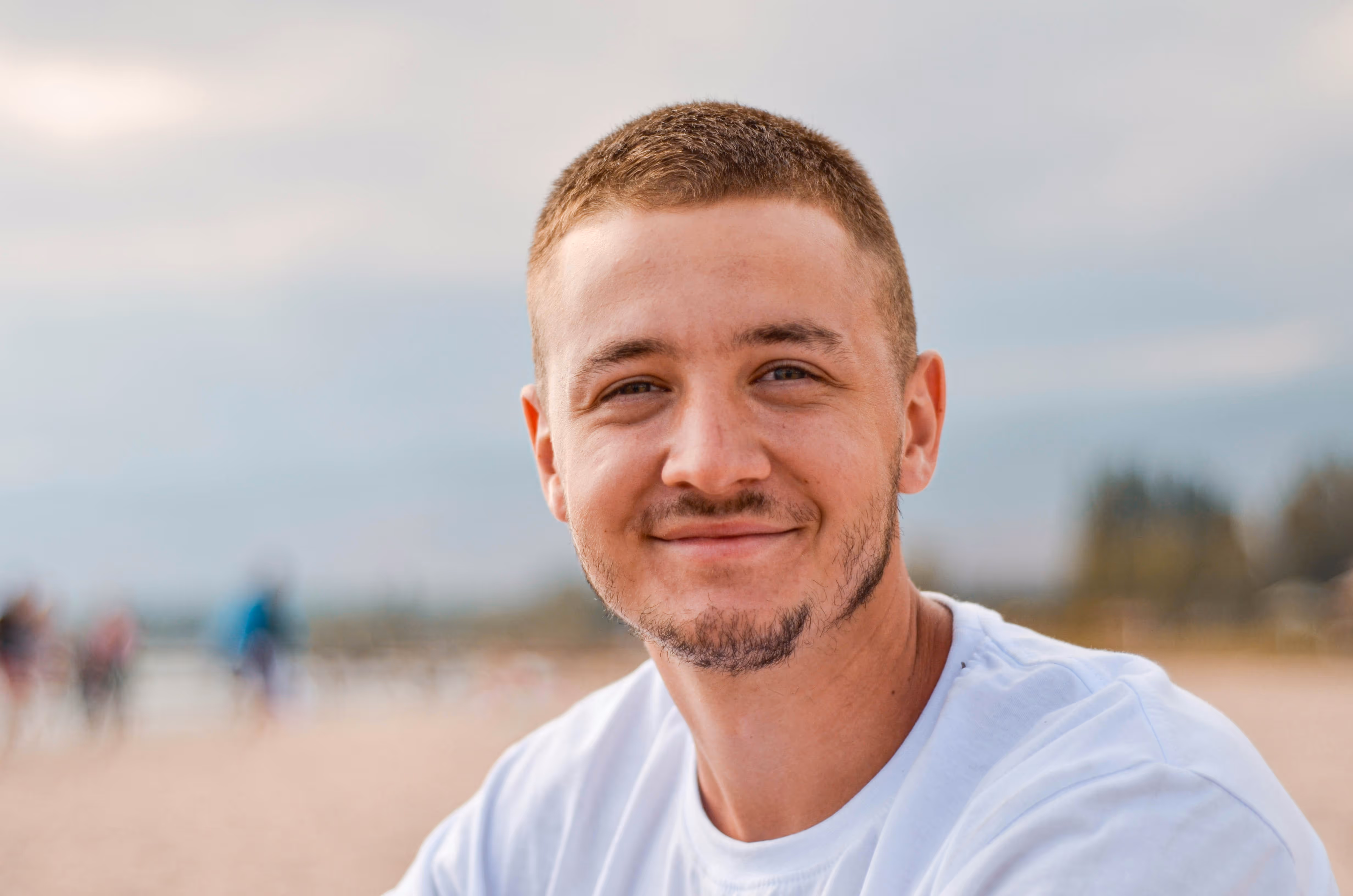 Smiling young man with short hair and light facial hair wearing a white shirt outdoors by the beach.