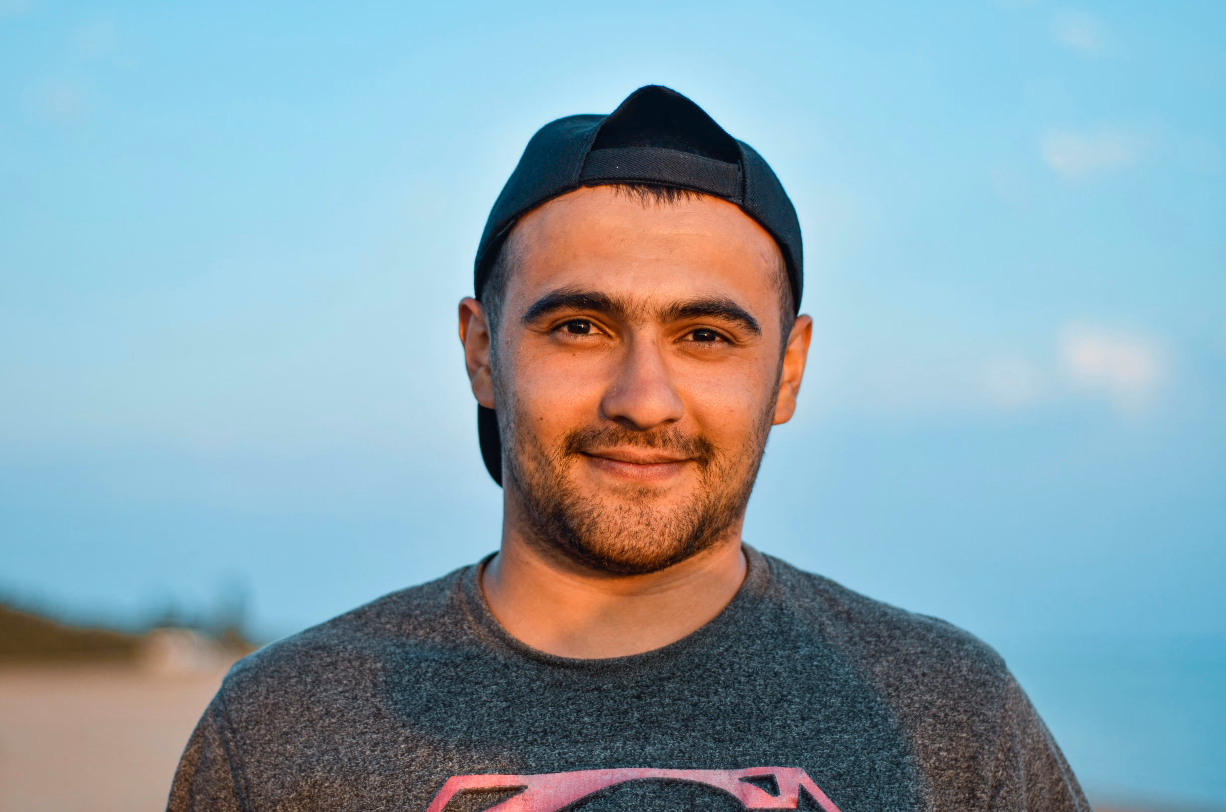 Smiling man wearing a backward black cap and gray Superman t-shirt outdoors at sunset.